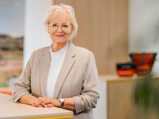 Anke Tammen with short blonde hair and glasses, wearing a beige blazer over a white turtleneck, standing in a bright office.
