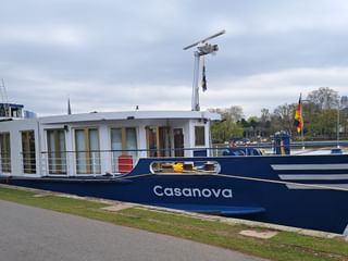 Blau-weißes Flusskreuzfahrtschiff MS Casanova am grasbewachsenen Flussufer unter bewölktem Himmel, mit Bäumen und Gebäuden im Hintergrund.