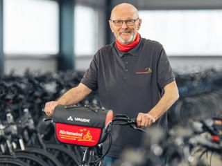 Juergen Bunge mit Brille und rotem Schal steht mit einem Fahrrad in einer großen Lagerhalle voller Fahrradreihen.