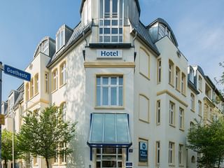 Weißes Eckgebäude mit markanter Turmspitze und blauem 'Hotel'-Schild. Hotel Geheimer Rat mit gelben Fensterrahmen und modernem Glaseingang.