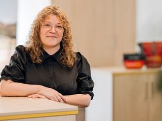 Marscha Büsing with curly blonde hair wearing a black blouse, sitting at a light wooden desk in a bright office with blurred background.