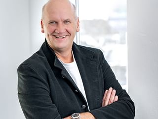 Andreas Bunge with arms crossed, wearing a dark blazer over a white shirt, smiling in a bright office with windows in the background.
