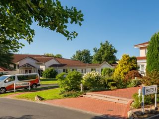 Weiße Hotelgebäude mit roten Ziegeldächern in Auefeld, umgeben von grünem Rasen und buntem Garten. Hotel-Shuttle vor dem Gebäude.