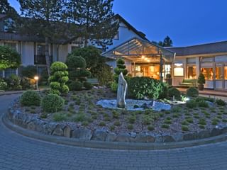 Landhotel Schnuck in der Dämmerung mit beleuchtetem Eingang und moderner Glasfassade. Runder Garten mit Skulptur und Zierpflanzen im Vordergrund.