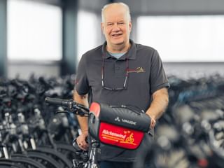 Carsten Rehbein steht mit einem Fahrrad in einer Lagerhalle voller Fahrradreihen. Er trägt ein graues Poloshirt und hält eine rote Tasche.