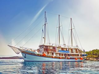 Weißes dreimastiges Segelschiff MS Sveti-Vid vor Anker in ruhigen blauen Gewässern nahe der bewaldeten kroatischen Küste unter bewölktem Himmel.