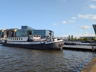 Weißes Passagierschiff MS Wending am Kai einer Uferpromenade mit modernen Amsterdamer Gebäuden und blauem Himmel im Hintergrund.
