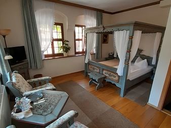 Geräumiges Hotelzimmer in Bautzen mit antikem blauen Himmelbett, blumig gepolsterten Sitzmöbeln, Holzboden und Bogenfenstern mit Vorhängen.