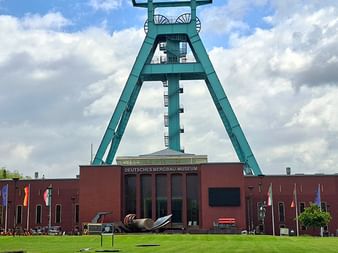 Ikonischer türkisfarbener Förderturm überragt das rote Backsteingebäude des Deutschen Bergbau-Museums in Bochum. Grüne Wiese im Vordergrund.