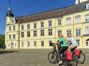 Zwei Radfahrer mit Tourenrädern und roten Packtaschen vor dem gelben barocken Oldenburger Schloss unter blauem Himmel.