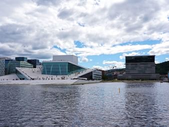 Osloer Opernhaus mit charakteristischem weißen Schrägdach am Fjord, mit modernen Bürogebäuden und Baukränen unter bewölktem Himmel.