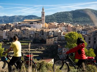 Zwei Radfahrer mit Rädern betrachten ein Dorf am Hang mit Kirchturm in der Region Valencia, Spanien. Berge erheben sich im Hintergrund.