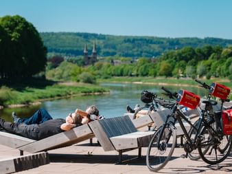 Zwei Radfahrer liegen auf Holzbänken neben der Weser mit ihren Tourenrädern in der Nähe. Kirchtürme sind in der Ferne sichtbar.