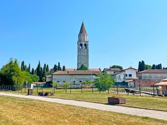 Hoher steinerner Glockenturm über historischen Gebäuden in Aquileia, Friaul. Grüne Wiese und Kiesweg im Vordergrund unter blauem Himmel.
