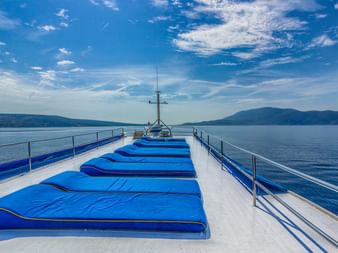 Sonnendeck des Schiffes MY Linda mit blauen gepolsterten Liegen auf weißem Deck, Metallgeländern und Küstengebirge unter blauem Himmel in Kroatien.