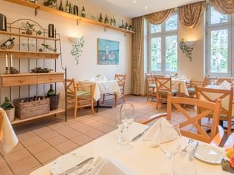 Heller Restaurantraum mit Holztischen, weißen Tischdecken und Stühlen mit Kreuzlehne. Große Fenster mit drapierten Vorhängen und Deko-Regalen.