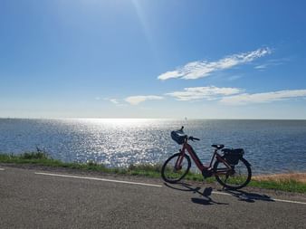 Rotes Fahrrad mit Taschen auf einem Weg am IJsselmeer unter blauem Himmel mit Sonnenlicht auf dem Wasser.