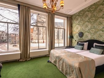 Elegantes Doppelzimmer mit zwei Betten, grünem Teppich, Ziertapete und großen Fenstern mit Blick auf einen Kanal in Deventer.