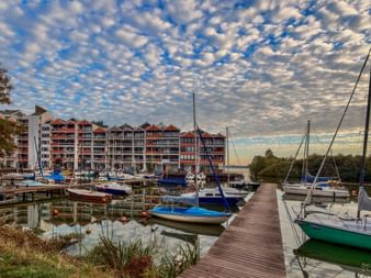 Hafen in Rastede mit Segelbooten am Holzsteg. Moderne Wohngebäude säumen das Ufer unter dramatischem Wolkenhimmel bei Sonnenuntergang.