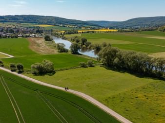 Luftaufnahme von Radfahrern auf einem Feldweg durch grüne Felder im Weserbergland. Die Weser schlängelt sich durch das Tal mit Hügeln.