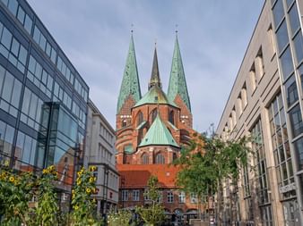 Rote Backsteinkirche mit grünen Kupfertürmen überragt moderne Gebäude in Lübecks Innenstadt, eingerahmt von zeitgenössischer Architektur.