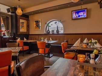 Gemütliche Hotelbar mit orangen Stühlen, Holztischen, braunen Sitzbänken und dekorativer Wandkunst. Ein TV-Bildschirm und Rundbogen sind sichtbar.