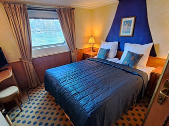 Schiffskabine auf der MS Casanova mit blauer Bettwäsche, blauer Akzentwand, Fenster mit Wasserblick, TV und gemustertem Teppich.