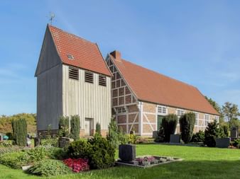 Kirche mit orangefarbenem Ziegeldach und hölzernem Glockenturm in Rastede, umgeben von Friedhof mit Grabsteinen und grüner Wiese unter blauem Himmel.