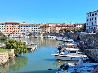 Malerischer Hafenkanal in der Toskana mit weißen Booten an Steinmauern, bunte historische Gebäude säumen das Ufer unter blauem Himmel.