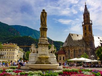 Steinbrunnen mit goldener Statue auf Bozens Hauptplatz, umgeben von bunten Blumen. Gotischer Dom mit verziertem Turm und Bergen im Hintergrund.