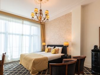 Elegantes Hotelzimmer im Huis Vermeer mit Doppelbett, Kronleuchter, großen Fenstern mit Vorhängen, gemustertem Teppich und dunklen Holzmöbeln.
