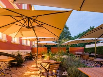 Außenterrasse im Parkhotel Jever mit gelben Sonnenschirmen, Holztischen und Stühlen, umgeben von Pflanzen und orangefarbenen Wänden unter Abendhimmel.