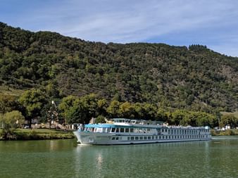 MS Switzerland Flusskreuzfahrtschiff auf ruhigem Wasser mit bewaldeten Bergen und kleinem Dorf im Hintergrund unter blauem Himmel.