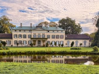 Schloss Rastede, ein klassizistisches Herrenhaus mit grünen Fensterläden, spiegelt sich in einem Teich. Das Gebäude ist von gepflegten Rasenflächen und alten Bäumen umgeben.