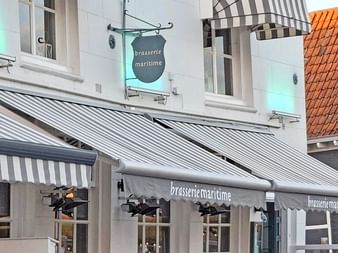 Fassade des Restaurants Brasserie Maritime in Zierikzee mit grau-weiß gestreiften Markisen, weißem Gebäude und sichtbarem orangefarbenem Ziegeldach.