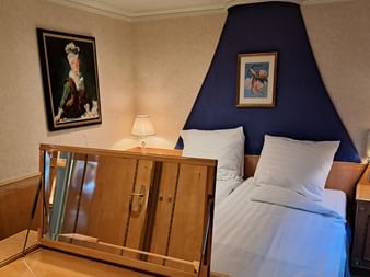 Kabineninterieur auf der MS Casanova mit Einzelbett, weißer Bettwäsche, blauem Stoffkopfteil, Holzmöbeln, gerahmter Kunst und Nachttischlampen.