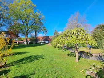 Weitläufiger Garten mit grünem Rasen, blühenden Sträuchern und Bäumen am Bösehof in Bad Bederkesa. Häuser mit roten Dächern im Hintergrund unter blauem Himmel.