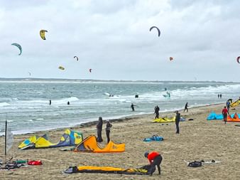 Strand bei Neeltje Jans mit Kitesurfern, bunten Drachen am Himmel, Ausrüstung im Sand und einer Windkraftanlage rechts.