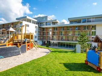 Modernes Alpenhotel mit Holzbalkonen und Kinderspielplatz mit bunten Klettergeräten, Rutschen und grüner Rasenfläche.