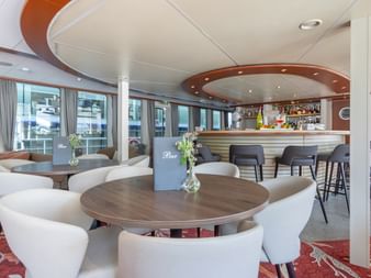 Modern ship lounge on MS Lisabelle with white chairs around round tables, curved bar with stools, large windows, and red patterned carpet.