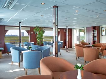 Modern ship library lounge on SE Manon with blue and orange armchairs, bookshelves, plants, and large windows overlooking the Danube river.