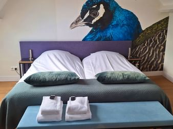 Boutique-Hotelzimmer am IJsselmeer mit Doppelbett, petrolgrüner Bettwäsche, weißen Handtüchern und großem Pfau-Foto über lila Kopfteil.