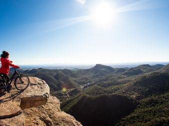 Zwei Radfahrer in roten Jacken auf Mountainbikes am felsigen Klippenrand mit Blick auf bewaldete Berge und Täler in der Region Valencia, Spanien.