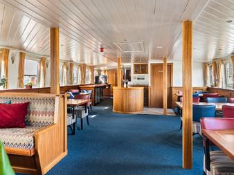 Geräumiges Restaurant an Bord der Floor van Aemstel mit Holztischen, bunten Sitzgelegenheiten, gemusterten Bänken und Fenstern an beiden Seiten.