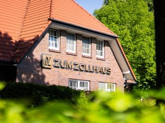 Rotes Backsteingebäude mit orangefarbenem Ziegeldach und 'Zum Zollhaus' Schriftzug an der Fassade, umgeben von grüner Vegetation.