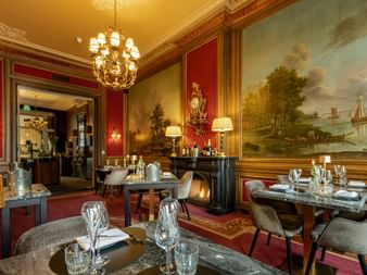 Elegantes Restaurant mit roten Wänden, prächtigem Kronleuchter und großem niederländischem Landschaftsgemälde. Tische mit Gläsern und grauen Stühlen.