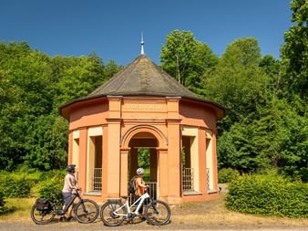 Zwei Radfahrer mit E-Bikes stehen neben einem runden orangefarbenen Pavillon mit Kegeldach in Birresborn. Das historische Lindenquelle-Gebäude ist von üppigem grünem Wald umgeben.