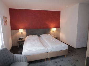 Modernes Hotelzimmer mit zwei Einzelbetten, roter Akzentwand, Holzkopfteil, Nachttischlampen, grauem Sessel und Fenster mit weißen Vorhängen.