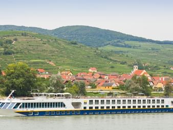 Flusskreuzfahrtschiff SE Manon auf der Donau in der Wachau. Dahinter ein Dorf mit roten Dächern und terrassierten Weinbergen an Hängen.