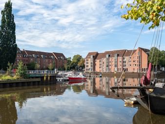 Kanal im Hafen Oldenburg mit traditionellen Segelbooten, modernen Wohngebäuden und Bäumen, die sich im ruhigen Wasser spiegeln.